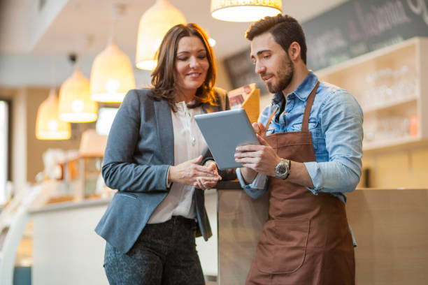 Man wearing apron and businesswomen looking at a digital tablet standing by bar counter in a restaurant. Owner and bar tender going through daily problems.
