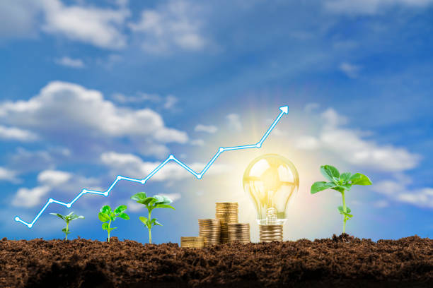 Close up view of a financial growth graph, coin stacks, plant pods and a glowing light bulb on soil shot against defocused blue sky with clouds background. Focus on foreground