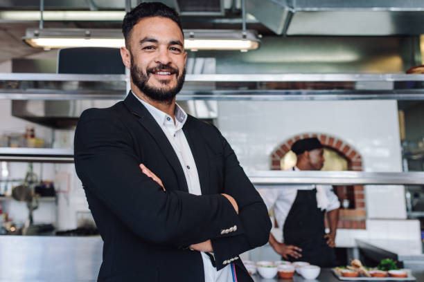 Portrait of young businessman standing in his restaurant with staff in kitchen. Proud restaurant owner standing with his arms crossed and looking at camera.